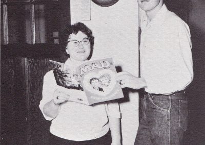 Female and male student standing in front of office both are holding a single issue of Mad Magazine. The boy is significantly taller and has his finger on the fire alarm lever.