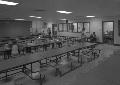 Black and white photo of Robertson Elementary. One table is being used by students. Teacher is helping two students near line.